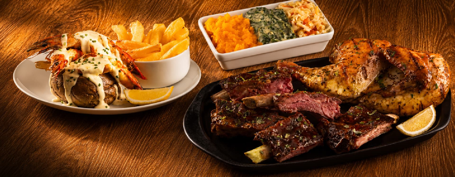 Mixed grill platter featuring barbecue pork ribs, roasted chicken, creamy garlic prawns on a baked potato, golden fries, mashed vegetables, creamed spinach, coleslaw, and lemon wedges, served on a rustic wooden table