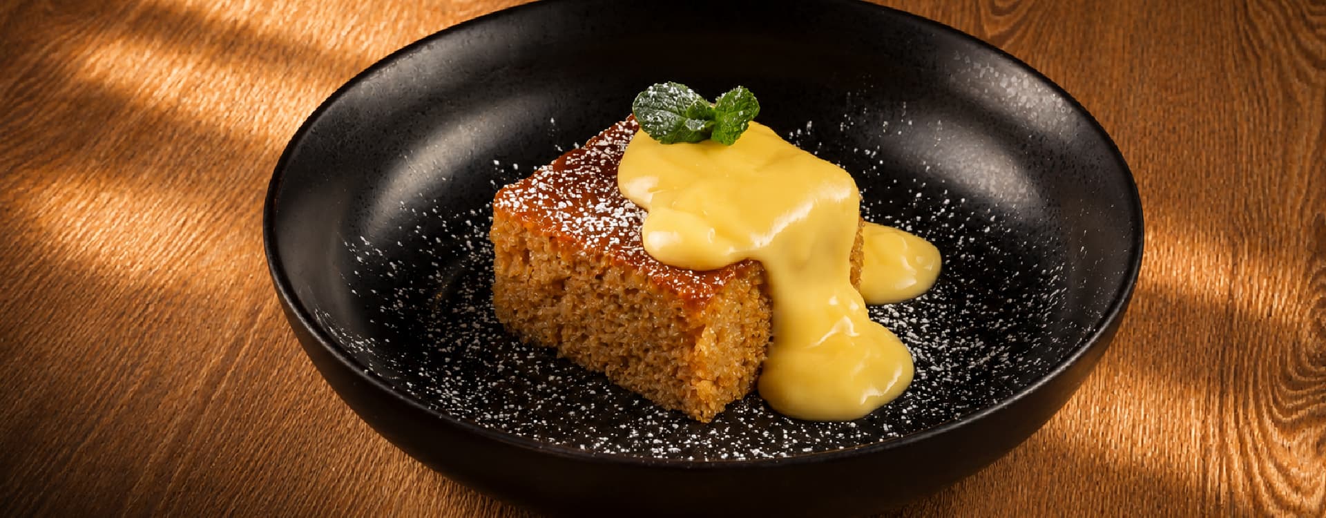 Traditional South African malva pudding topped with rich vanilla custard and a fresh mint garnish, dusted with powdered sugar and served in a black bowl