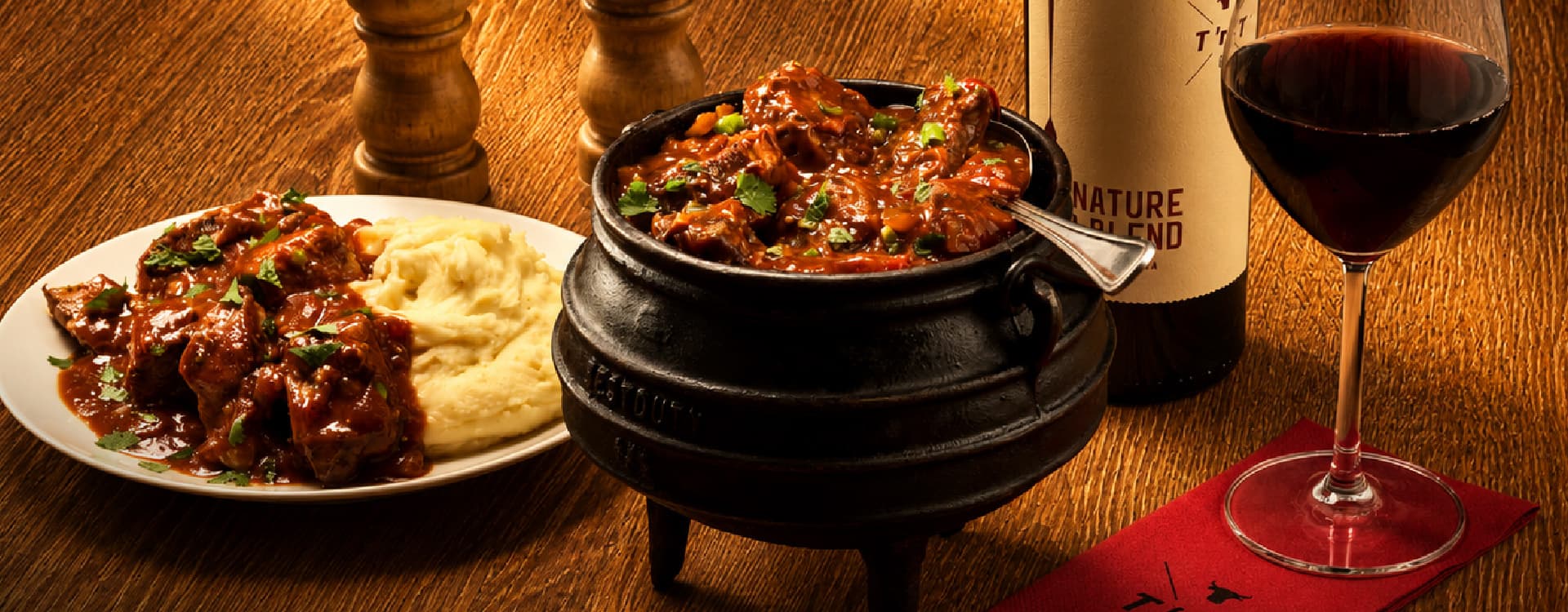 Traditional South African beef potjie simmered in a rich tomato-based gravy with herbs, served in a cast-iron pot alongside creamy mashed potatoes. A bottle of red wine and a glass of red wine complete the rustic dining setting