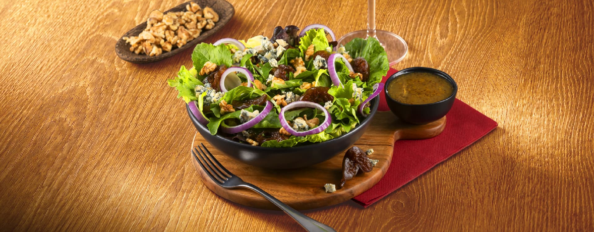 Fresh green salad with lettuce, blue cheese crumbles, red onion rings, walnuts, and dried fruit, served in a black bowl with vinaigrette dressing on a wooden serving board