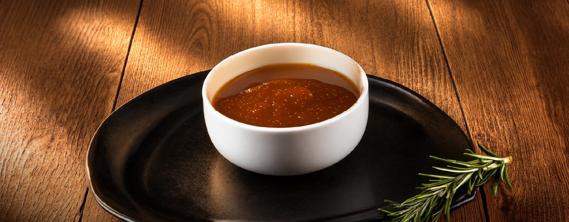 Small white ramekin filled with glossy sauce, presented on a black plate with a sprig of fresh rosemary on a rustic wooden tabletop