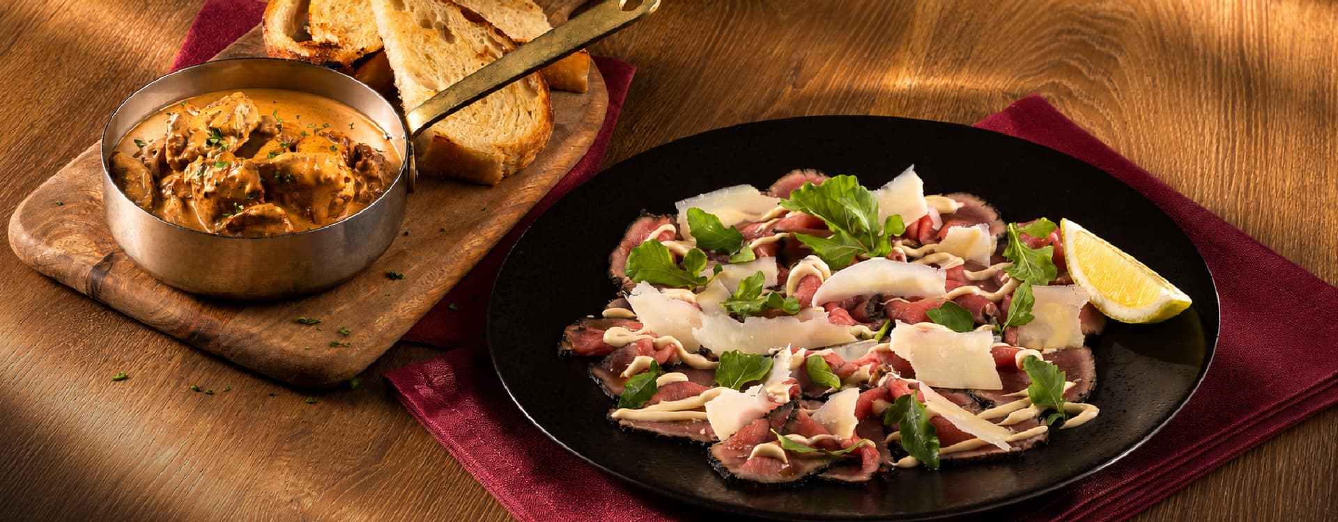 Thinly sliced beef carpaccio topped with shaved Parmesan cheese, rocket leaves, and creamy dressing, served with a lemon wedge. Accompanied by a rich creamy mushroom dish and toasted artisan bread on a rustic wooden table