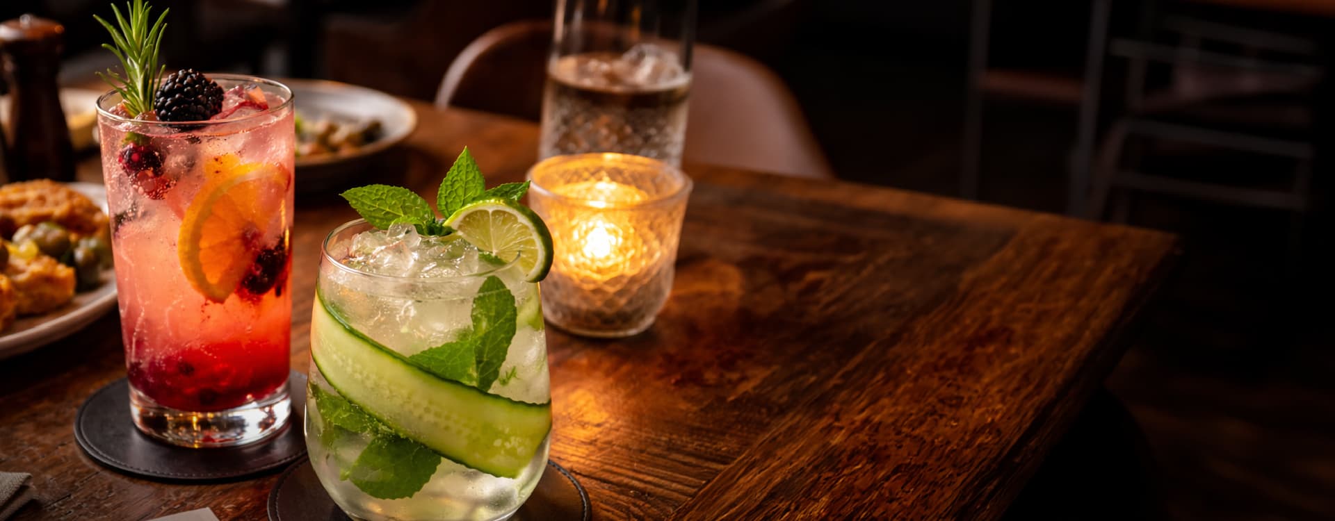 Refreshing cocktails featuring cucumber, lime, mint, berries, and citrus served over ice on a candlelit restaurant table