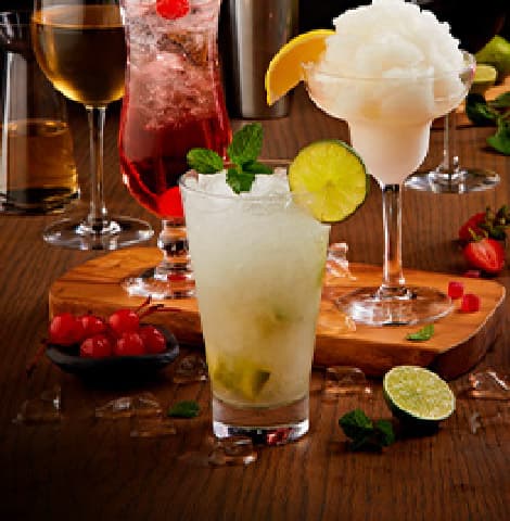 Handcrafted cocktail selection featuring a mojito, berry cocktail, frozen margarita, white wine, and fresh fruit garnishes displayed on a wooden bar table