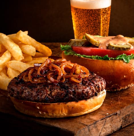 Open gourmet beef burger with caramelized onions, lettuce, tomato, pickles, and creamy sauce served with thick-cut fries and a glass of beer on a rustic wooden table