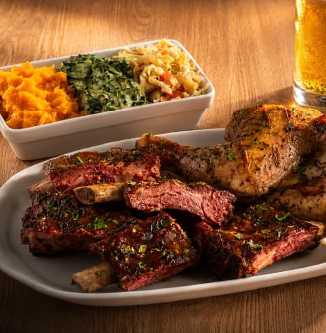 Restaurant-style mixed grill platter featuring barbecue pork ribs and herb-roasted chicken, served with mashed vegetables, creamed spinach, coleslaw, and a cold beer in the background