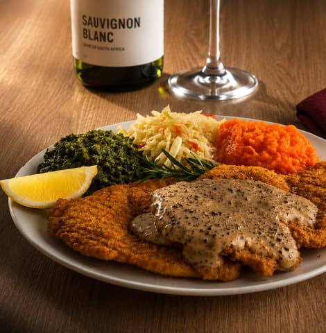 Crumbed schnitzel topped with creamy pepper sauce, served with sautéed spinach, coleslaw, and a lemon wedge on a white plate. A bottle labeled “Sauvignon Blanc” and a wine glass sit in the background