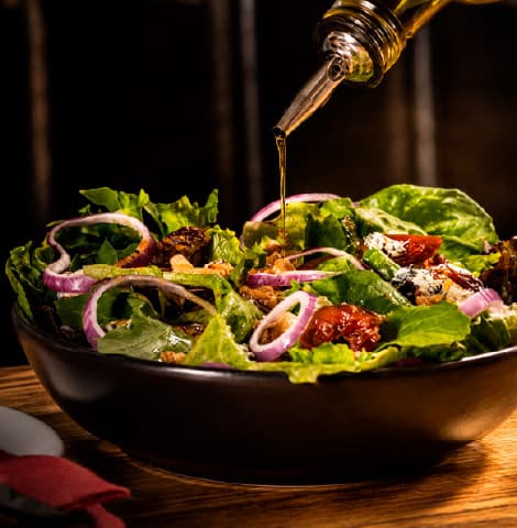 Fresh garden salad with crisp lettuce, red onion rings, walnuts, dried fruit, and blue cheese crumbles, drizzled with olive oil from a bottle above