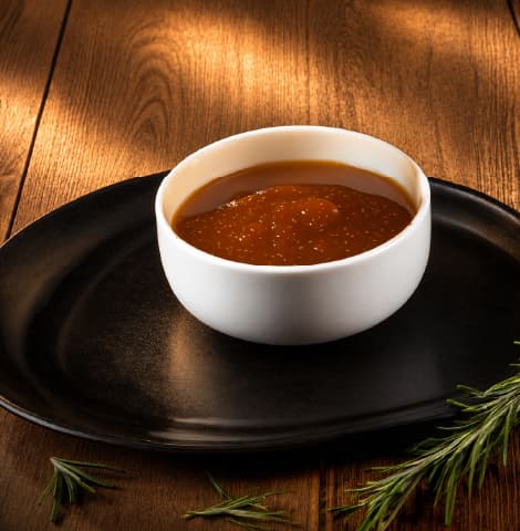 Small white ramekin filled with glossy sauce, presented on a black plate with a sprig of fresh rosemary on a rustic wooden tabletop