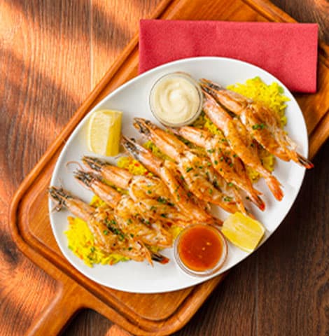 Grilled prawns covered in a creamy white sauce and garnished with chopped chives, served on a baked potato with thick-cut fries and lemon wedges. A glass of white wine is visible in the background