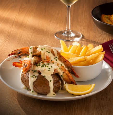 Grilled prawns covered in a creamy white sauce and garnished with chopped chives, served on a baked potato with thick-cut fries and lemon wedges. A glass of white wine is visible in the background