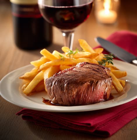 Juicy grilled beef steak served with golden French fries on a white plate, accompanied by a glass of red wine and a wine bottle in the background