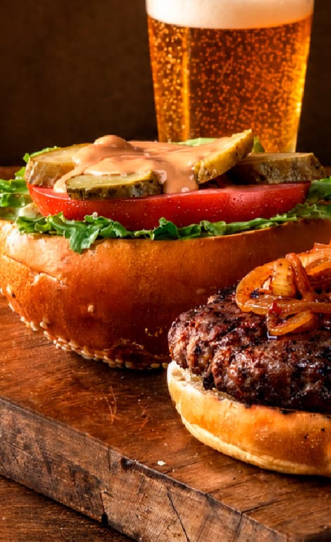 Open gourmet beef burger with caramelized onions, lettuce, tomato, pickles, and creamy sauce served with thick-cut fries and a glass of beer on a rustic wooden table