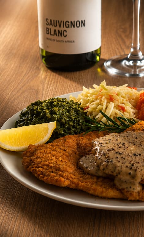 Crumbed schnitzel topped with creamy pepper sauce, served with sautéed spinach, coleslaw, and a lemon wedge on a white plate. A bottle labeled “Sauvignon Blanc” and a wine glass sit in the background