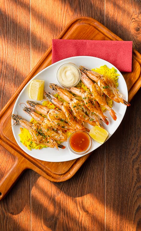 Grilled prawns covered in a creamy white sauce and garnished with chopped chives, served on a baked potato with thick-cut fries and lemon wedges. A glass of white wine is visible in the background