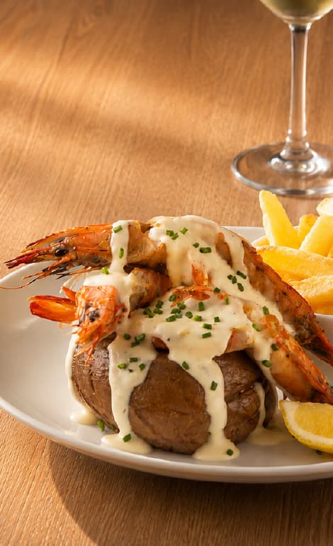 Grilled prawns covered in a creamy white sauce and garnished with chopped chives, served on a baked potato with thick-cut fries and lemon wedges. A glass of white wine is visible in the background