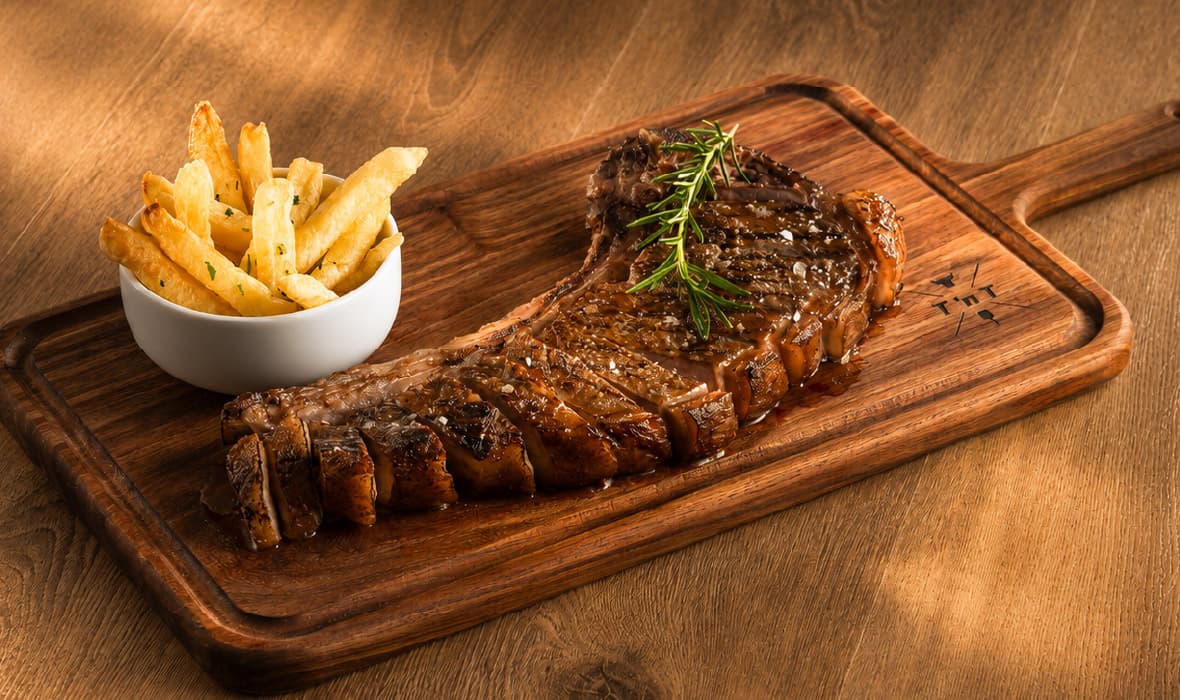 Sliced ribeye steak glazed with sauce and garnished with fresh rosemary, served on a wooden serving board with a bowl of golden fries