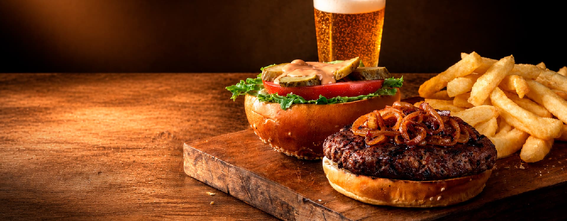 Open gourmet beef burger with caramelized onions, lettuce, tomato, pickles, and creamy sauce served with thick-cut fries and a glass of beer on a rustic wooden table