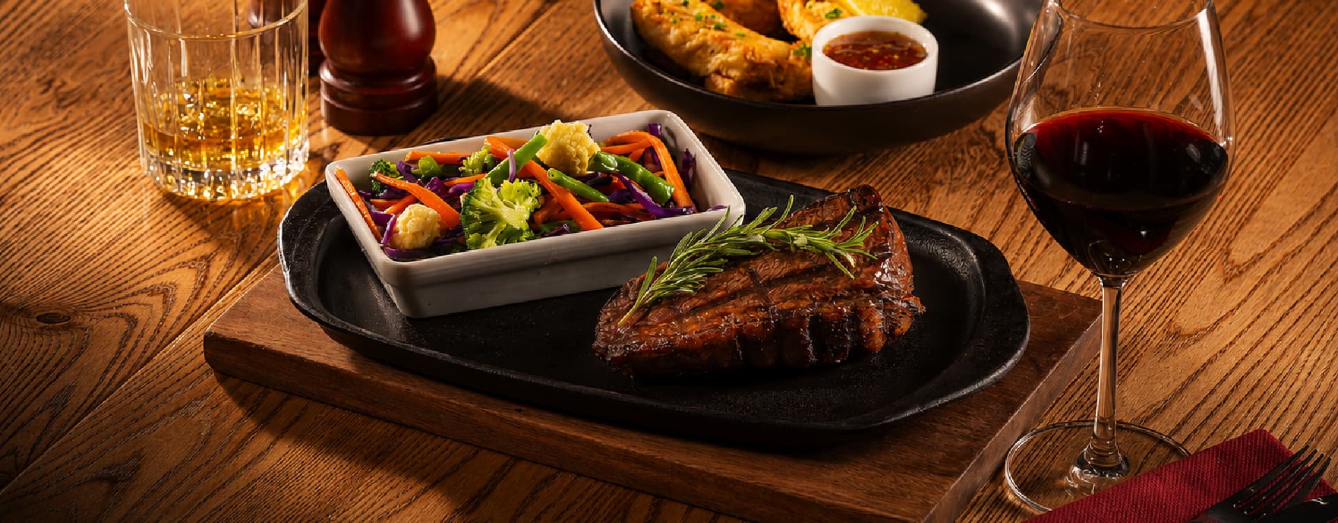 Char-grilled steak garnished with fresh rosemary, served with mixed vegetables on a sizzling black platter. A glass of red wine, whiskey tumbler, and appetizer plate are arranged on a rustic wooden dining table