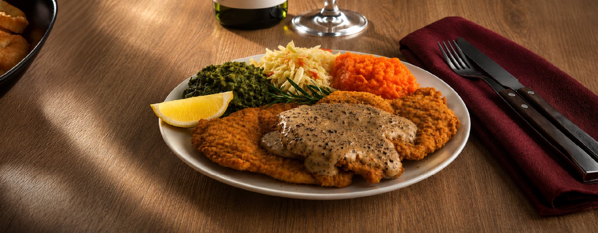 Crumbed schnitzel topped with creamy pepper sauce, served with sautéed spinach, coleslaw, and a lemon wedge on a white plate. A bottle of wine background