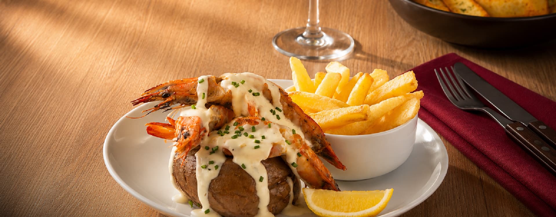 Grilled prawns topped with creamy garlic sauce and fresh chives, served on a baked potato with golden fries and a lemon wedge on a white plate