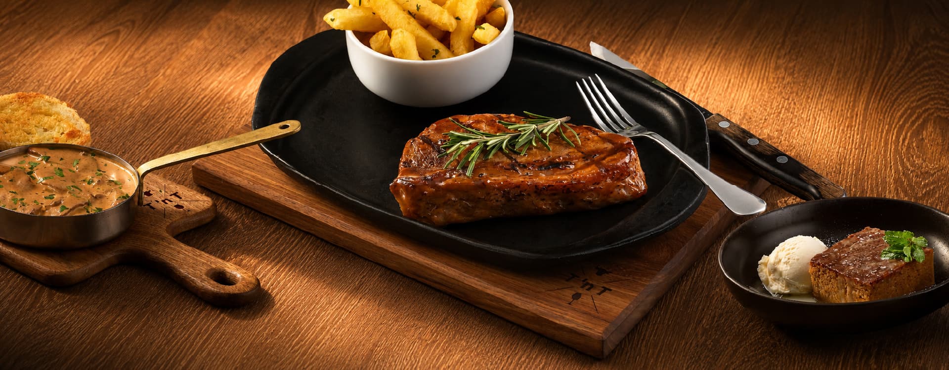 Char-grilled steak garnished with fresh rosemary, served on a sizzling platter with fries and mushroom sauce. A dessert of cake and vanilla ice cream is presented alongside on a rustic wooden table