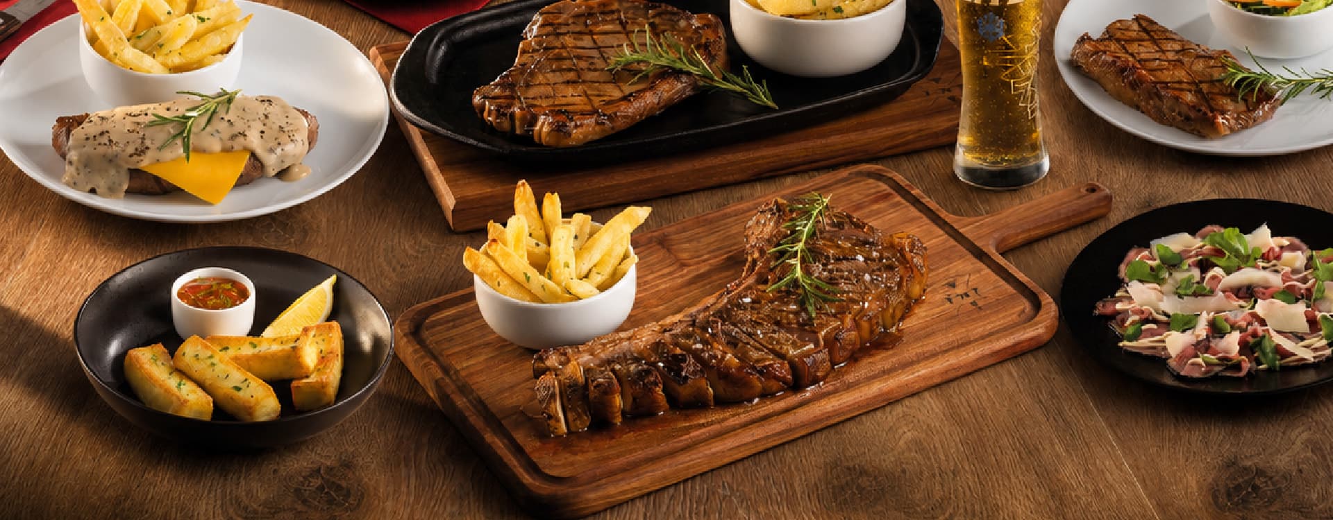 Assortment of premium steak dishes including grilled ribeye, T-bone steak, pepper steak, garlic bread, fries, salad, and beverages, arranged on a wooden table for a restaurant menu showcase
