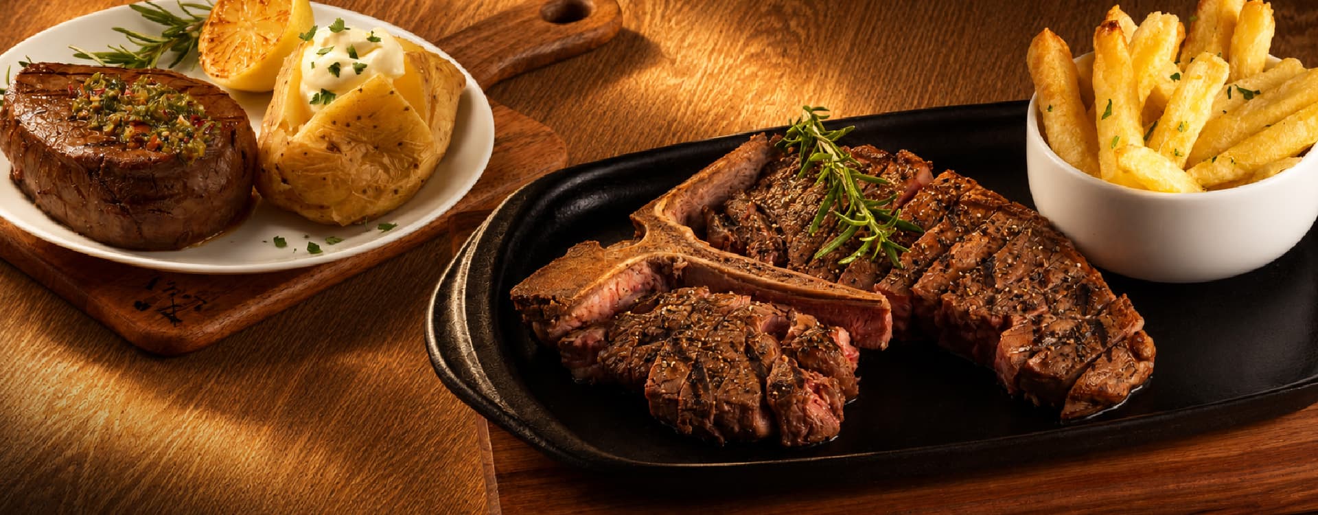 Juicy sliced T-bone steak seasoned with herbs and served on a sizzling platter with thick-cut fries. A grilled fillet steak, baked potato topped with sour cream, and grilled lemon are displayed alongside on a rustic wooden table