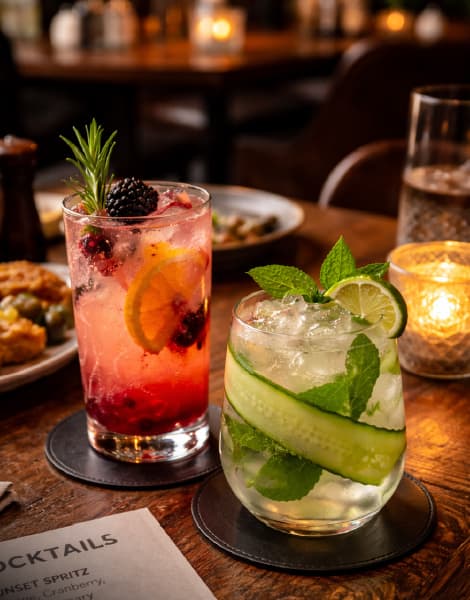Assorted handcrafted cocktails including a mojito, berry spritzer, frozen cocktail, and wine, garnished with lime, mint, cherries, and strawberries on a rustic wooden bar